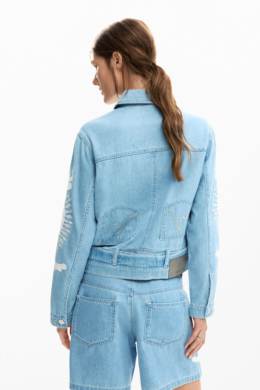 Back view of Woman wearing a light blue washed jean jacket with with embroidered detail all over