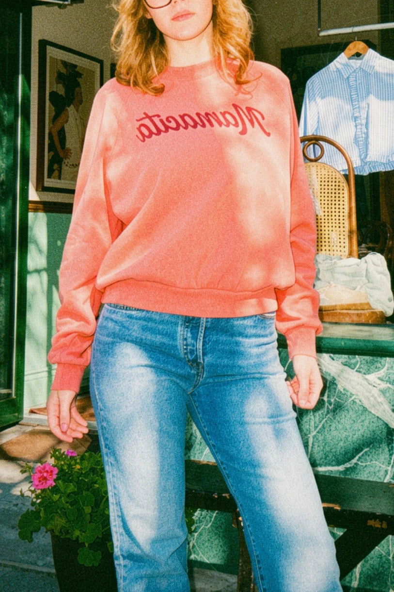 A woman wearing a coral pink crewneck sweatshirt with 'Mamawata' embroidered in red across the chest, paired with light-wash blue jeans, standing in front of a shop window.
