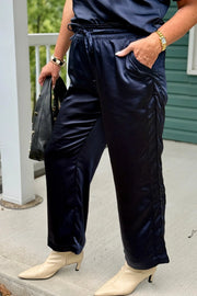 A person wearing shiny navy wide-leg pants with side pockets and a drawstring waist, paired with cream pointed-toe heeled boots, standing on a porch.