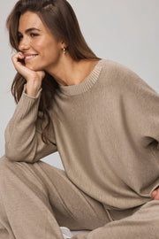 Woman wearing a beige sweater and pants sitting against a plain background