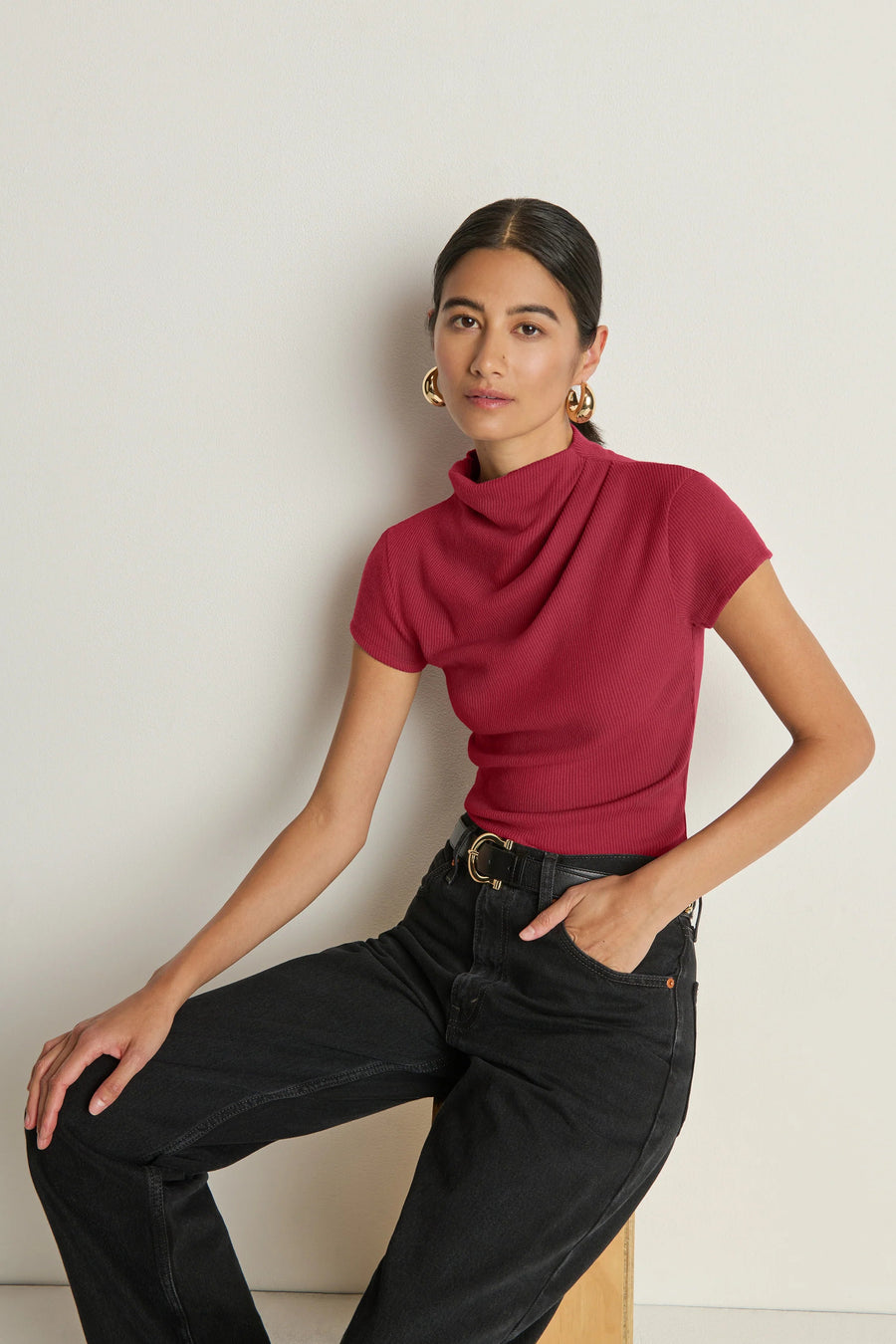 Woman wearing a red drape-neck top and black pants against a white wall.