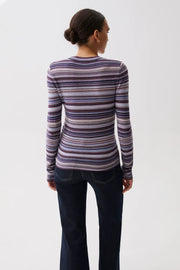 A woman modeling a long-sleeve crewneck knit top with horizontal stripes in purple, blue, pink, and beige, paired with dark jeans against a plain light gray studio background.