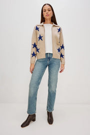 Woman wearing a beige cardigan with blue star patterns, white shirt, blue jeans, and brown boots on a white background.