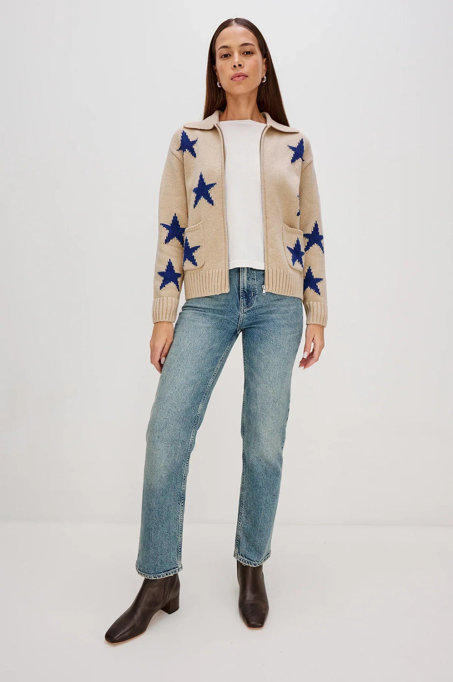 Woman wearing a beige cardigan with blue star patterns, white shirt, blue jeans, and brown boots on a white background.