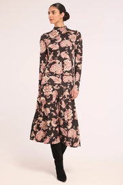 A woman modeling a long-sleeved midi dress with a black base and a pink floral paisley pattern, paired with black boots, against a plain light background.