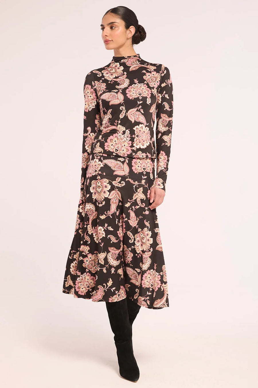 A woman modeling a long-sleeved midi dress with a black base and a pink floral paisley pattern, paired with black boots, against a plain light background.