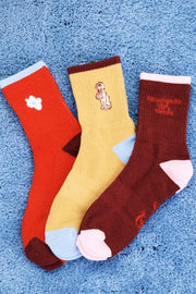Three pairs of crew socks in red, mustard yellow, and dark brown. Each sock features a different embroidered character or graphic on the front.