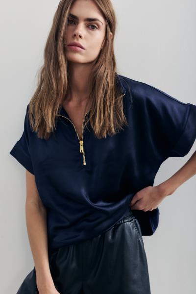 A woman wearing a navy blue satin short-sleeve top with a gold zipper at the collar. 
