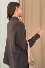 Woman wearing a dark brown sweater holding a beige handbag against a neutral background