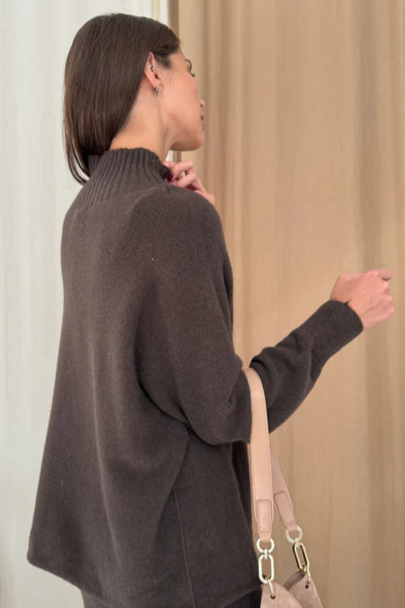 Woman wearing a dark brown sweater holding a beige handbag against a neutral background
