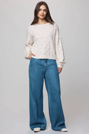 Woman wearing a cream sweater with cable knit pattern and pearl detailing all over paired with blue jeans