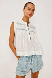 Woman wearing a flowy sleeveless white top with blue embroidery 
