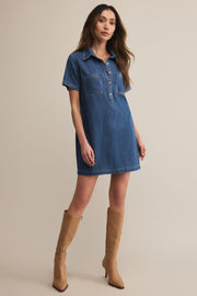 Woman wearing a blue denim dress with short sleeves and button details, paired with tan knee-high boots on a beige background.
