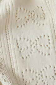 Woman wearing a cream cardigan with buttons on the front, scalloped trim and openwork detailing, fabric detail view