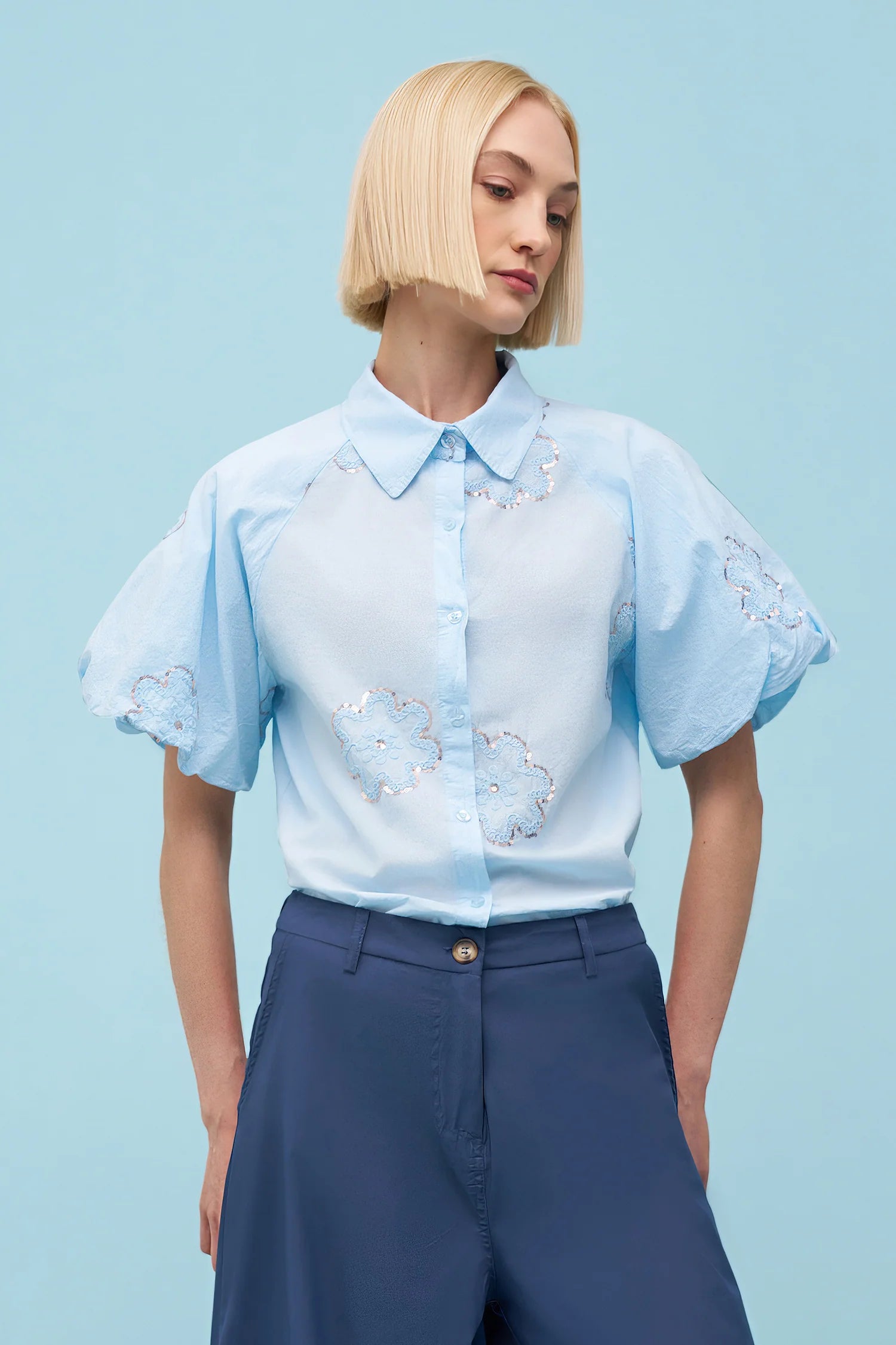Woman wearing a light blue short-sleeve button-up blouse with floral embroidery and a pointed collar, styled with navy high-waisted trousers against a matching blue background.
