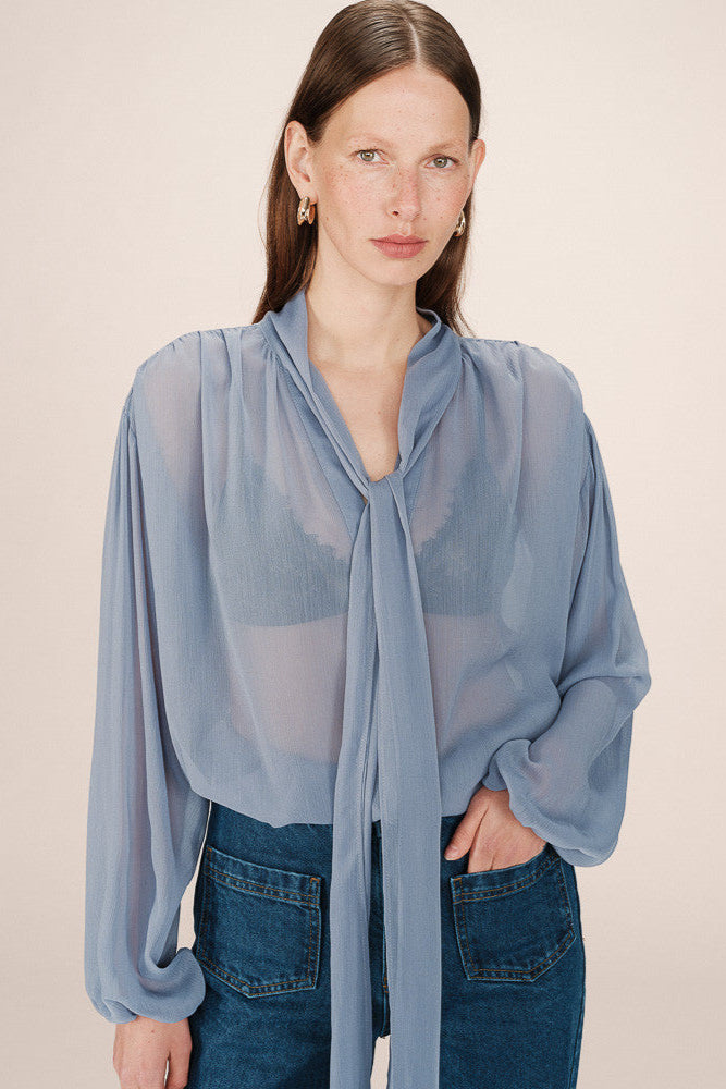 Front view of a woman wearing a sheer light blue blouse with a tie detail in the shoulders and  tie up neck 