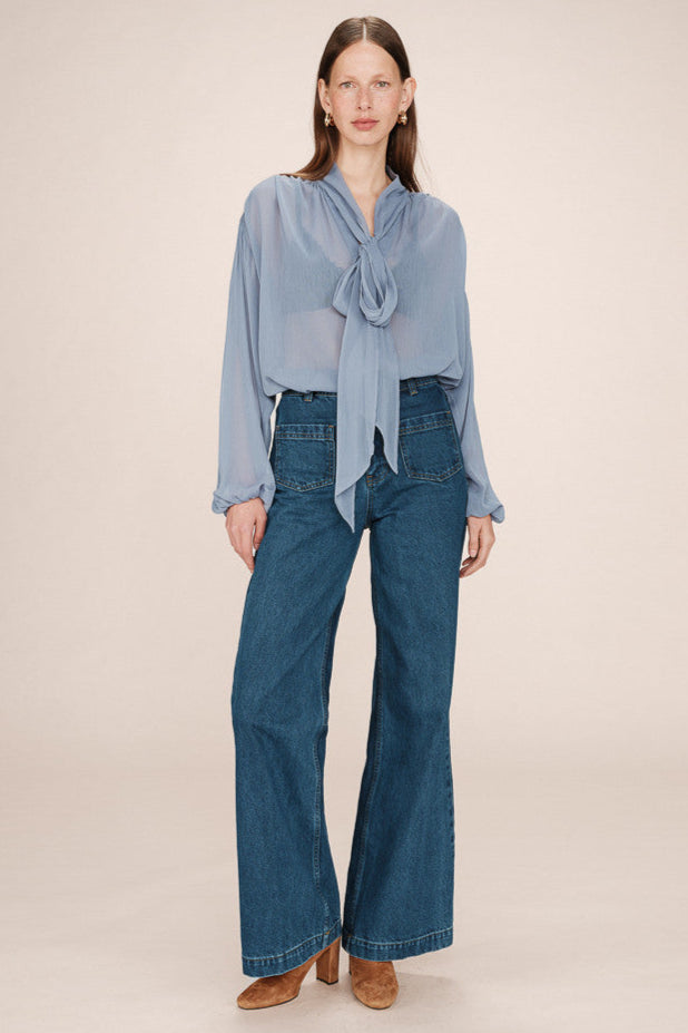 Front view of a woman wearing a sheer light blue blouse with a tie detail in the shoulders and  tie up neck with jeans