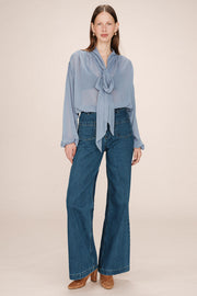 Front view of a woman wearing a sheer light blue blouse with a tie detail in the shoulders and  tie up neck with jeans