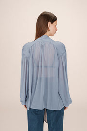 Back view of a woman wearing a sheer light blue blouse with a tie detail in the shoulders and  tie up neck 