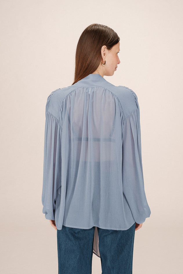 Back view of a woman wearing a sheer light blue blouse with a tie detail in the shoulders and  tie up neck 