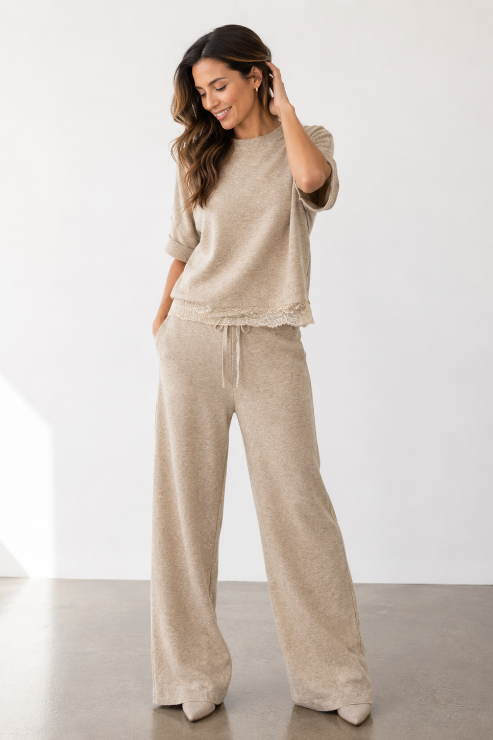 A woman modeling a matching beige knit lounge set consisting of a short-sleeve top with a ruffled hem and wide-leg drawstring pants