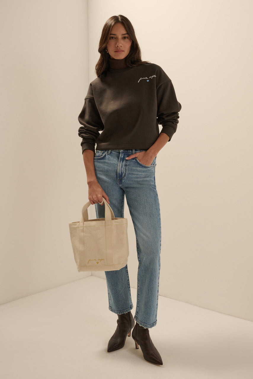 A woman stands in a minimalist studio setting wearing a dark brown mock-neck sweatshirt