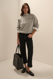 A woman stands in a minimalist studio setting wearing a light gray crewneck sweatshirt with a small embroidered logo on the chest,