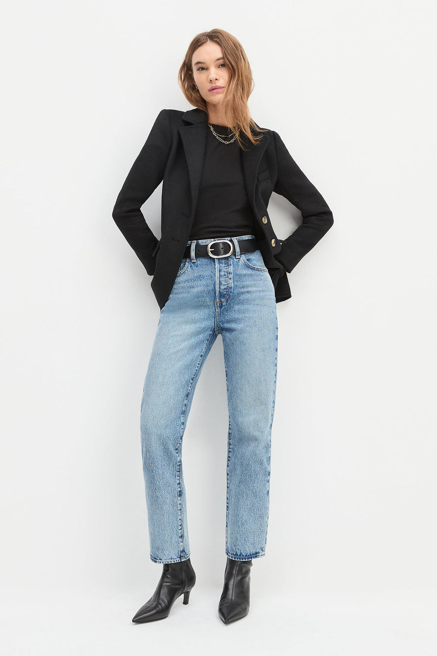 Woman wearing a black blazer, black top, light blue jeans, and black boots on a white background