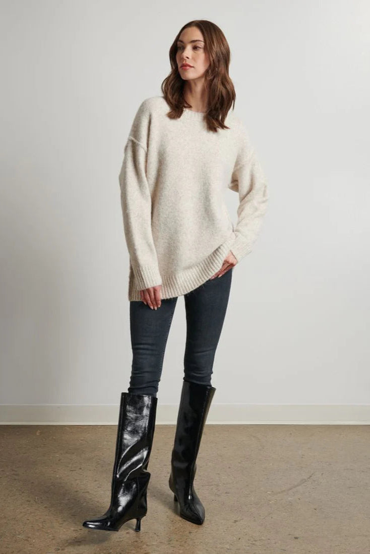 Woman wearing a cream sweater, dark jeans, and black boots against a plain background