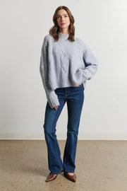 Woman wearing a light blue sweater and blue jeans standing against a plain white wall.