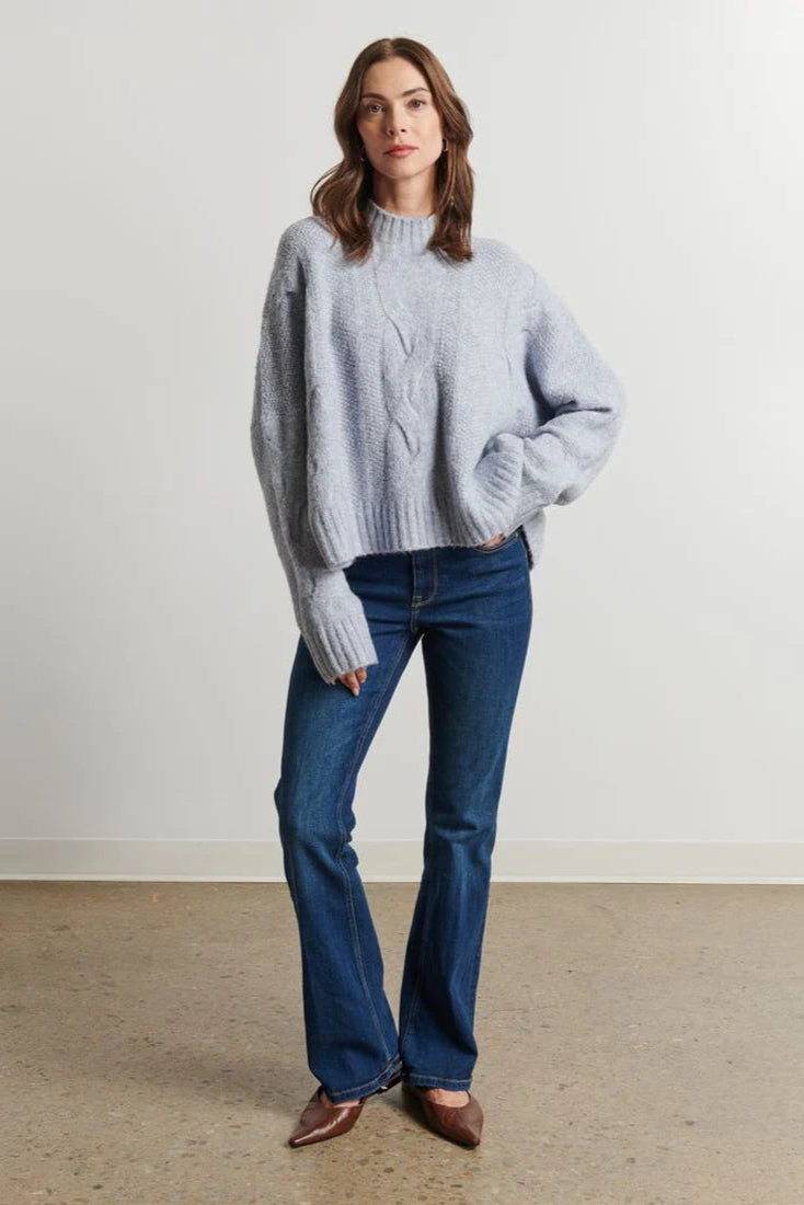 Woman wearing a light blue sweater and blue jeans standing against a plain white wall.