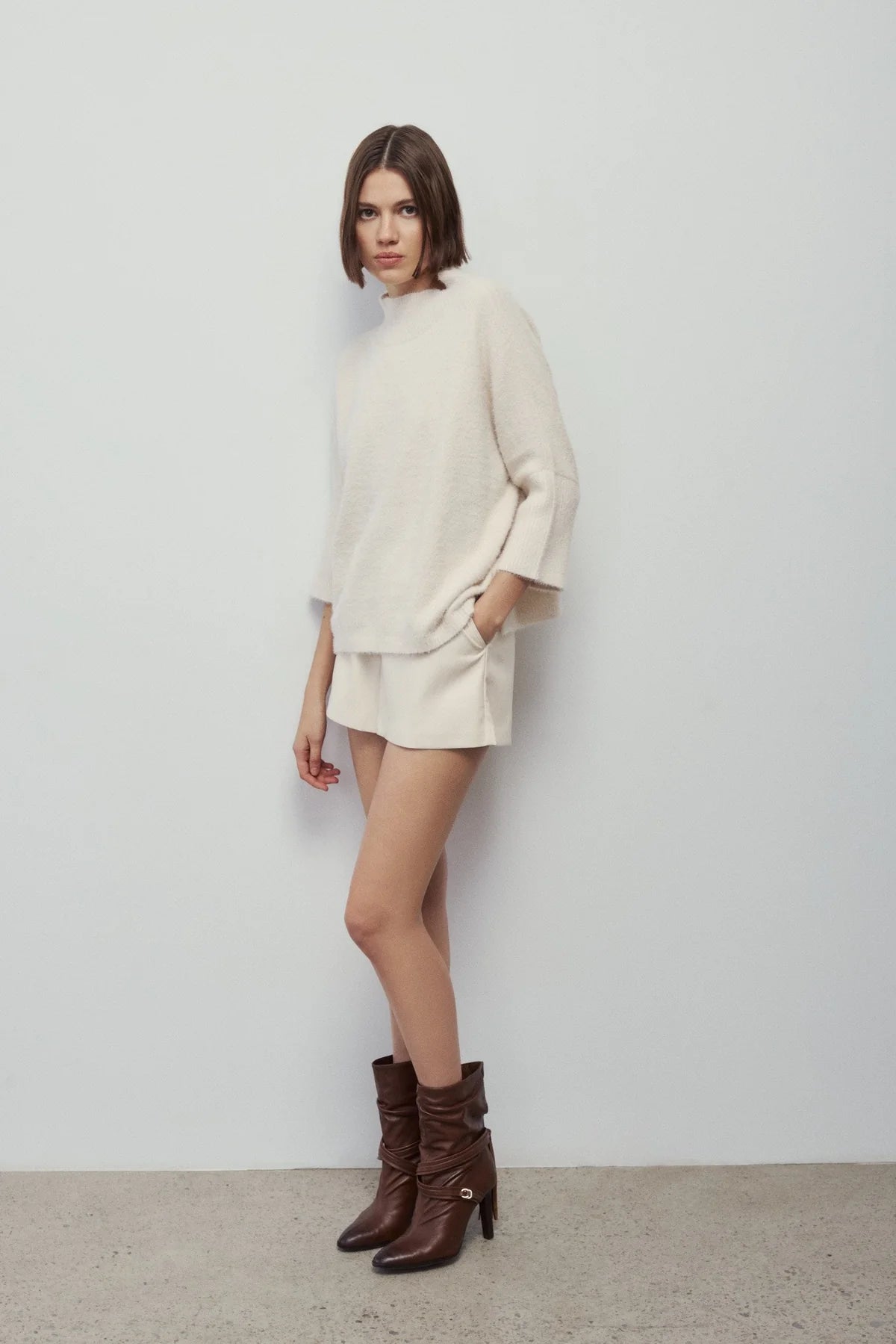 A woman modeling a cream-colored knit sweater and matching shorts set, paired with dark brown ankle boots, standing against a plain light gray wall.