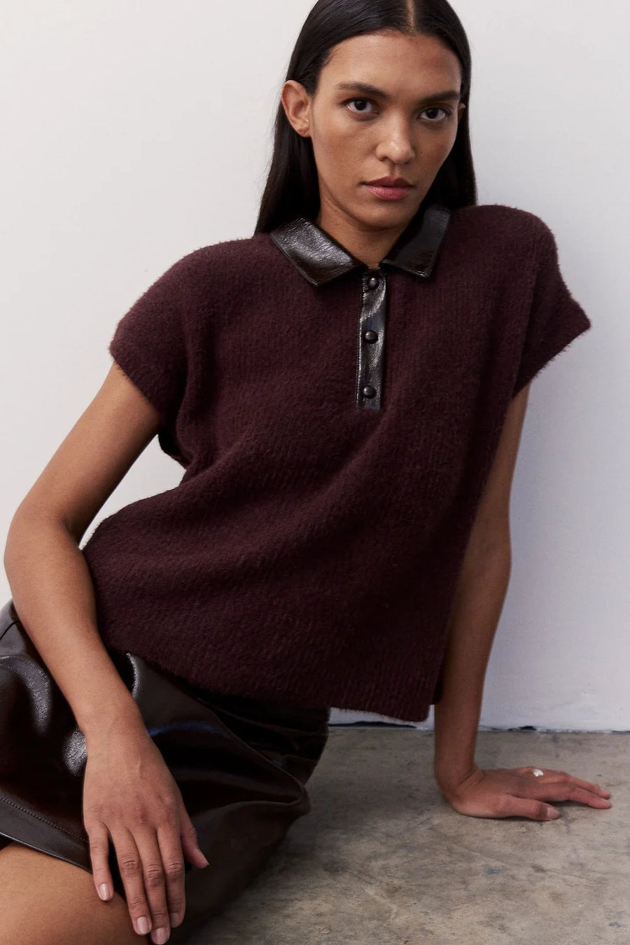 A woman wearing a short-sleeve knit polo top in deep burgundy with a dark brown collar and button placket, paired with a dark brown skirt, posing against a light gray wall.
