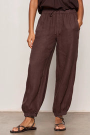 Woman wearing rich chocolate brown jogger-style pants with a relaxed fit. Features an elastic drawstring waistband, side pockets, and elasticized ankle cuffs. Crafted from a lightweight woven fabric with an easy, comfortable silhouette, front