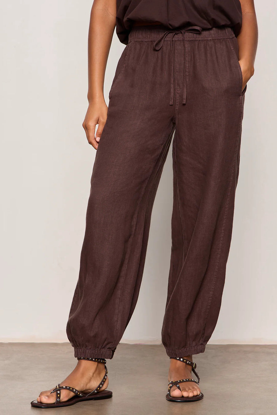 Woman wearing rich chocolate brown jogger-style pants with a relaxed fit. Features an elastic drawstring waistband, side pockets, and elasticized ankle cuffs. Crafted from a lightweight woven fabric with an easy, comfortable silhouette, front