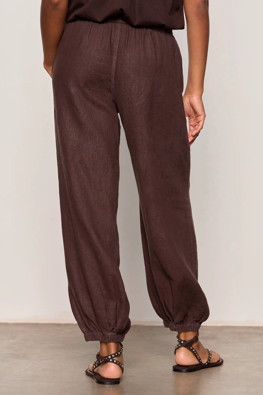 Woman wearing rich chocolate brown jogger-style pants with a relaxed fit. Features an elastic drawstring waistband, side pockets, and elasticized ankle cuffs. Crafted from a lightweight woven fabric with an easy, comfortable silhouette, back
