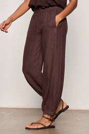 Woman wearing rich chocolate brown jogger-style pants with a relaxed fit. Features an elastic drawstring waistband, side pockets, and elasticized ankle cuffs. Crafted from a lightweight woven fabric with an easy, comfortable silhouette, side