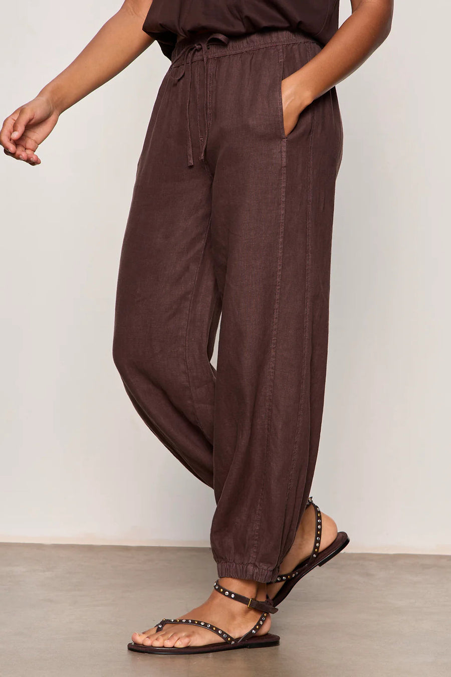 Woman wearing rich chocolate brown jogger-style pants with a relaxed fit. Features an elastic drawstring waistband, side pockets, and elasticized ankle cuffs. Crafted from a lightweight woven fabric with an easy, comfortable silhouette, side