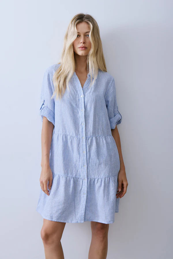 Woman wearing a light blue and white pinstripe shirt dress with a relaxed, tiered silhouette. Features a button-front closure, soft collar, rolled three-quarter sleeves with tab details, and a knee-grazing hemline. Designed in a lightweight woven fabric with subtle texture and effortless drape, front