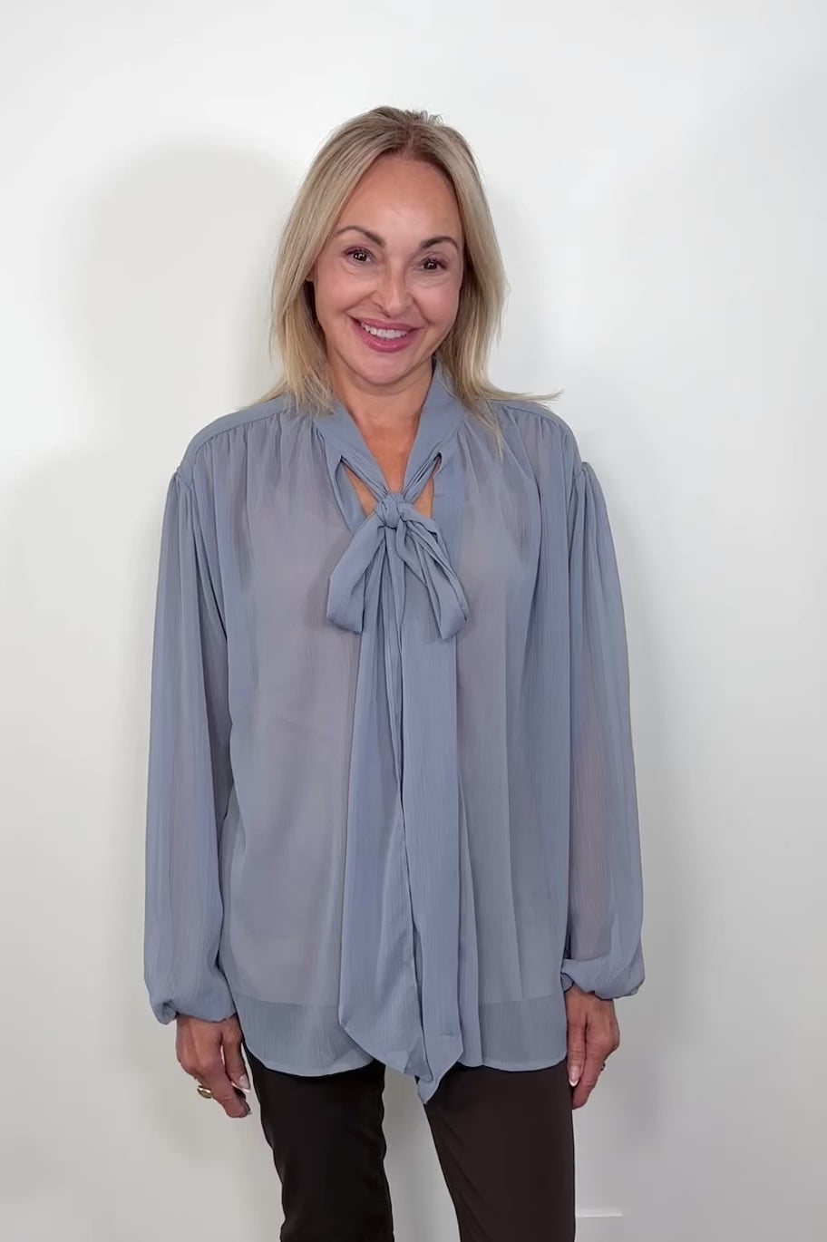 Video of woman wearing a thin blue blouse with a bow untucked turning around to show the back