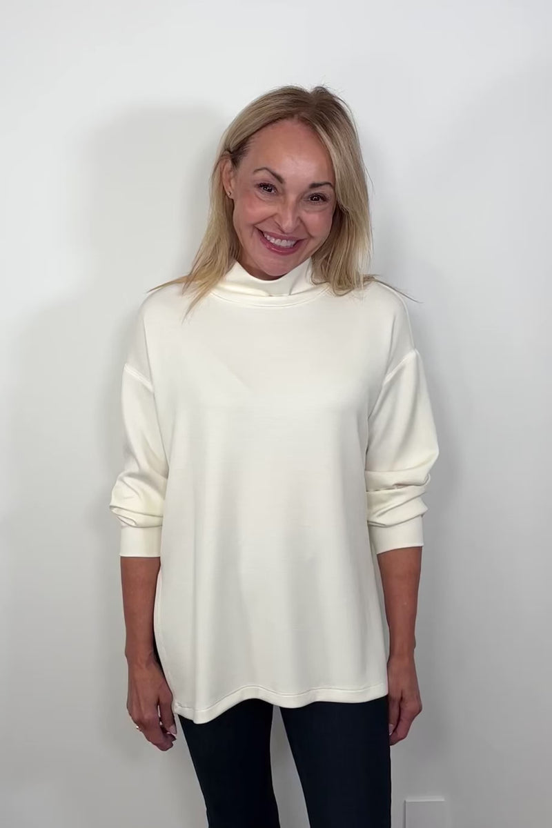 Video of a woman wearing a white boxy turtleneck top turning around to show the back