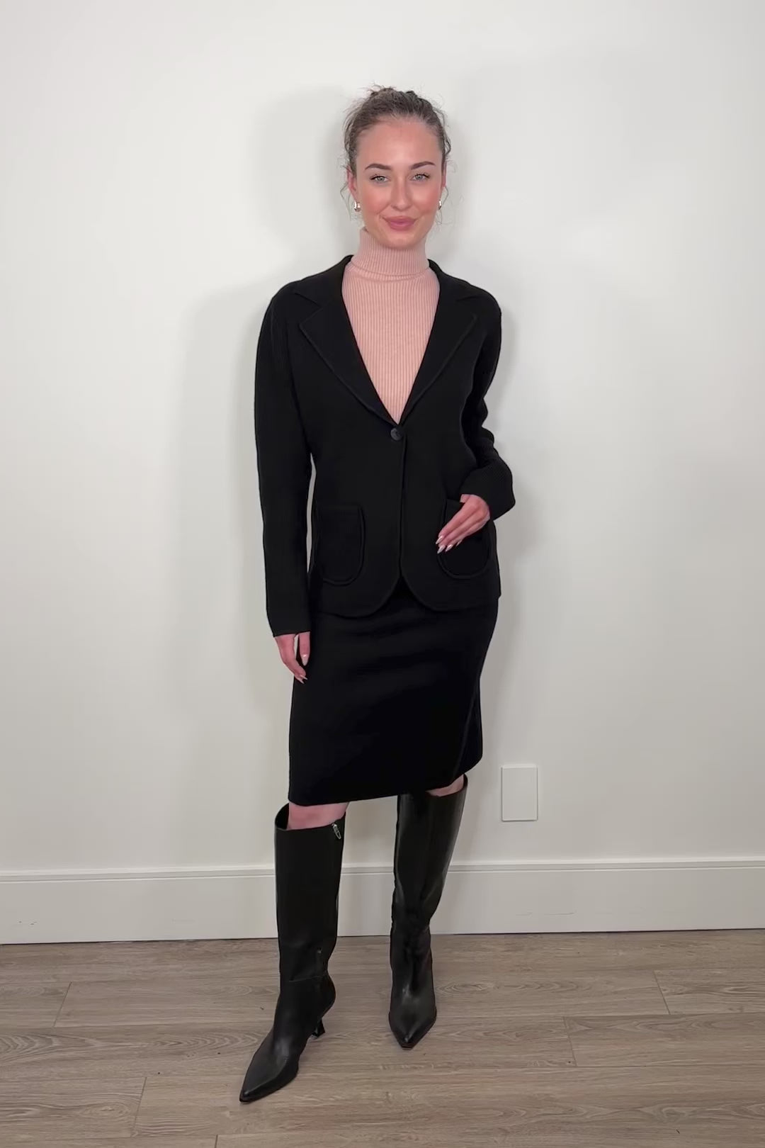 Video of woman wearing a matching black knitted blazer and skirt with tall boots and a pink turtleneck tank top