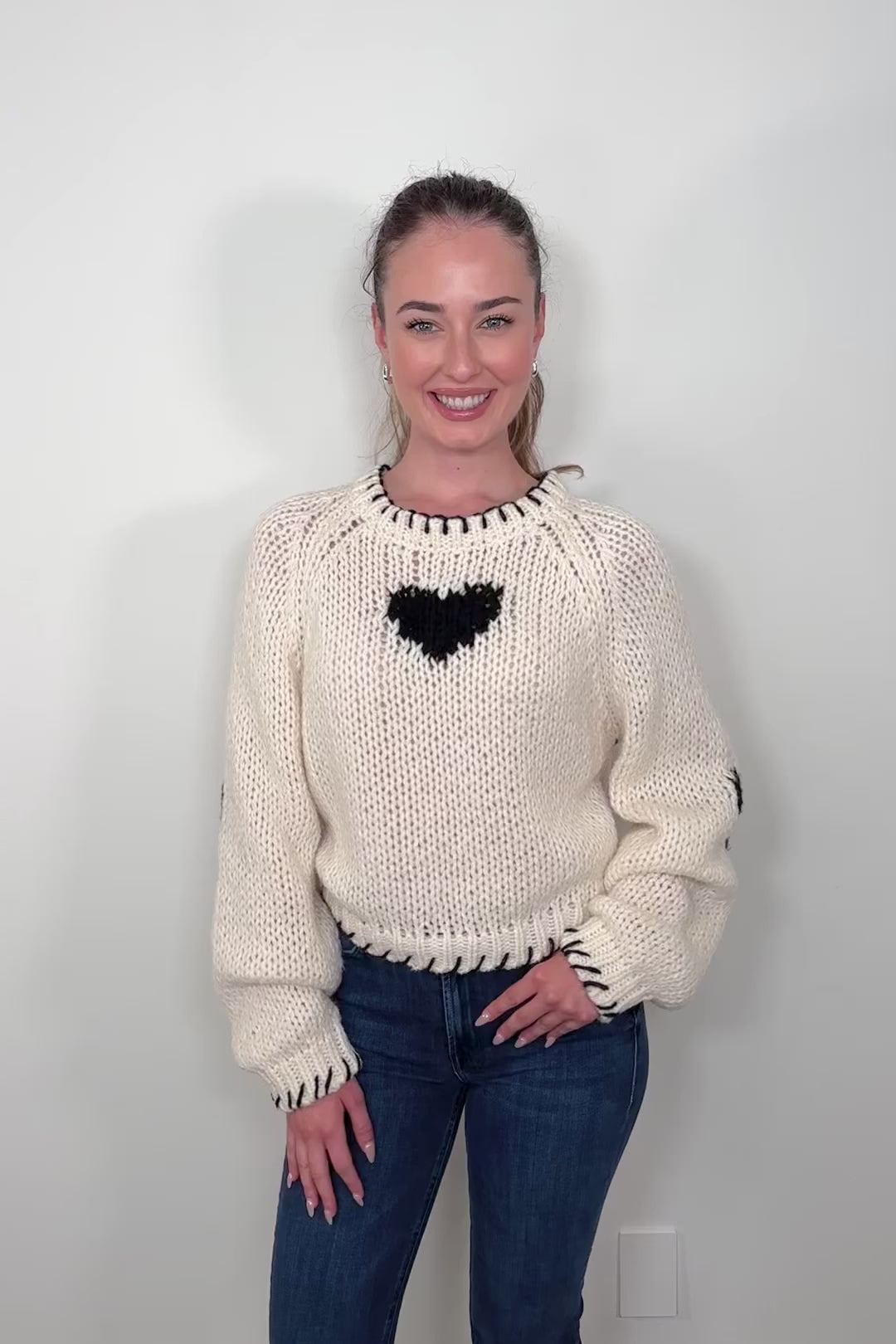 Video of a person wearing a cream-colored sweater with a black heart design and whip-stitch detailing on the collar