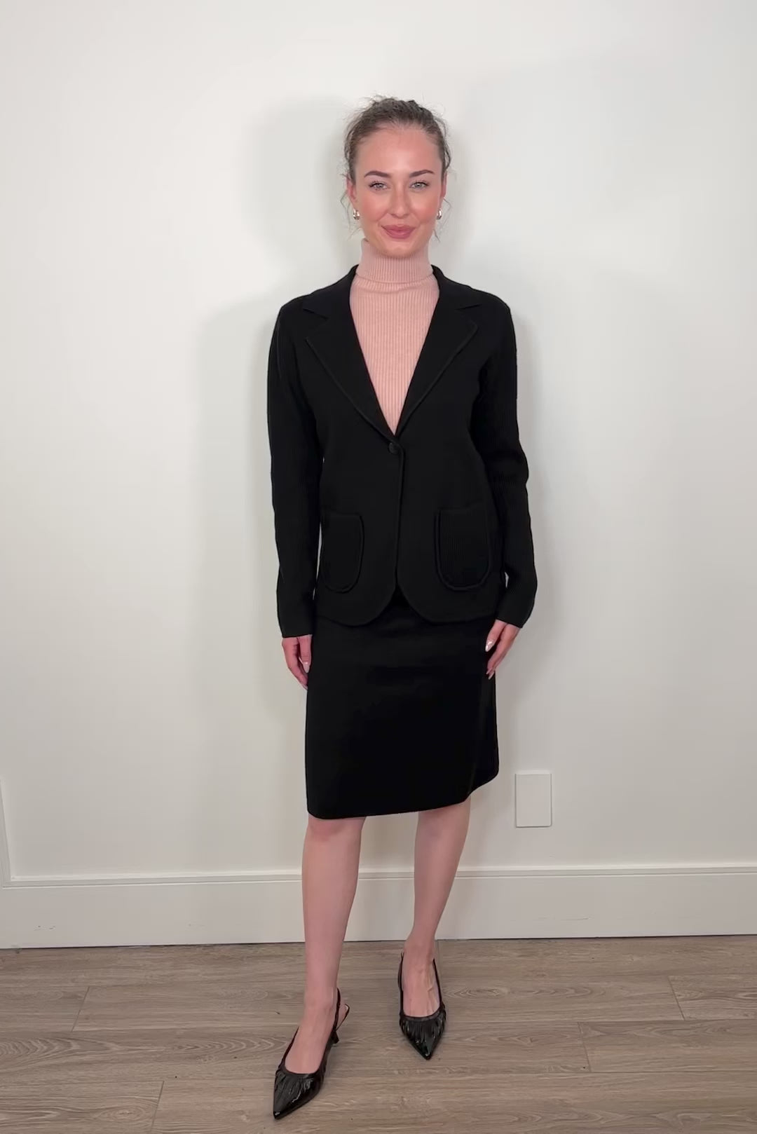 Video of woman wearing a matching black knitted blazer and skirt with heels and a pink turtleneck tank top