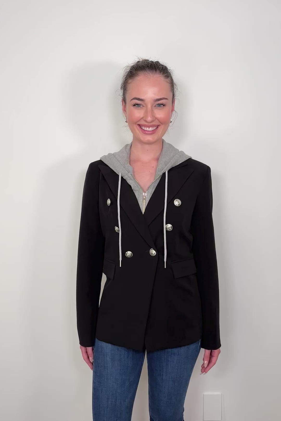 Video of woman wearing a black jacket that includes a detachable gray hoodie against a plain background