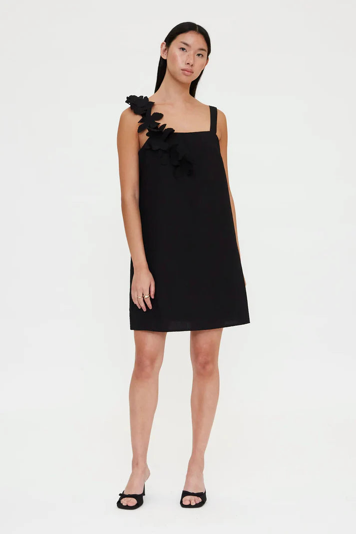 Woman wearing a black sleeveless mini dress with floral appliqué detailing on one shoulder, front