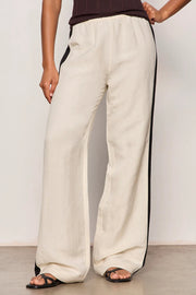 Woman wearing lightweight sand-colored wide-leg pants with contrasting black side stripe detailing. Features a comfortable pull-on elastic waistband, relaxed fit through the leg, and a fluid full-length silhouette. Designed with side pockets and a soft drape for effortless everyday wear, front
