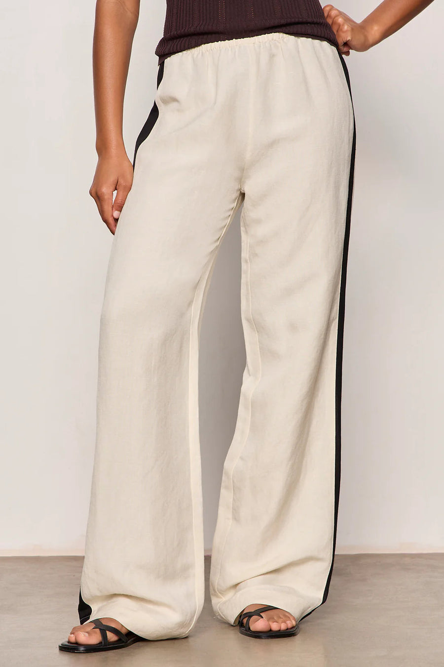 Woman wearing lightweight sand-colored wide-leg pants with contrasting black side stripe detailing. Features a comfortable pull-on elastic waistband, relaxed fit through the leg, and a fluid full-length silhouette. Designed with side pockets and a soft drape for effortless everyday wear, front