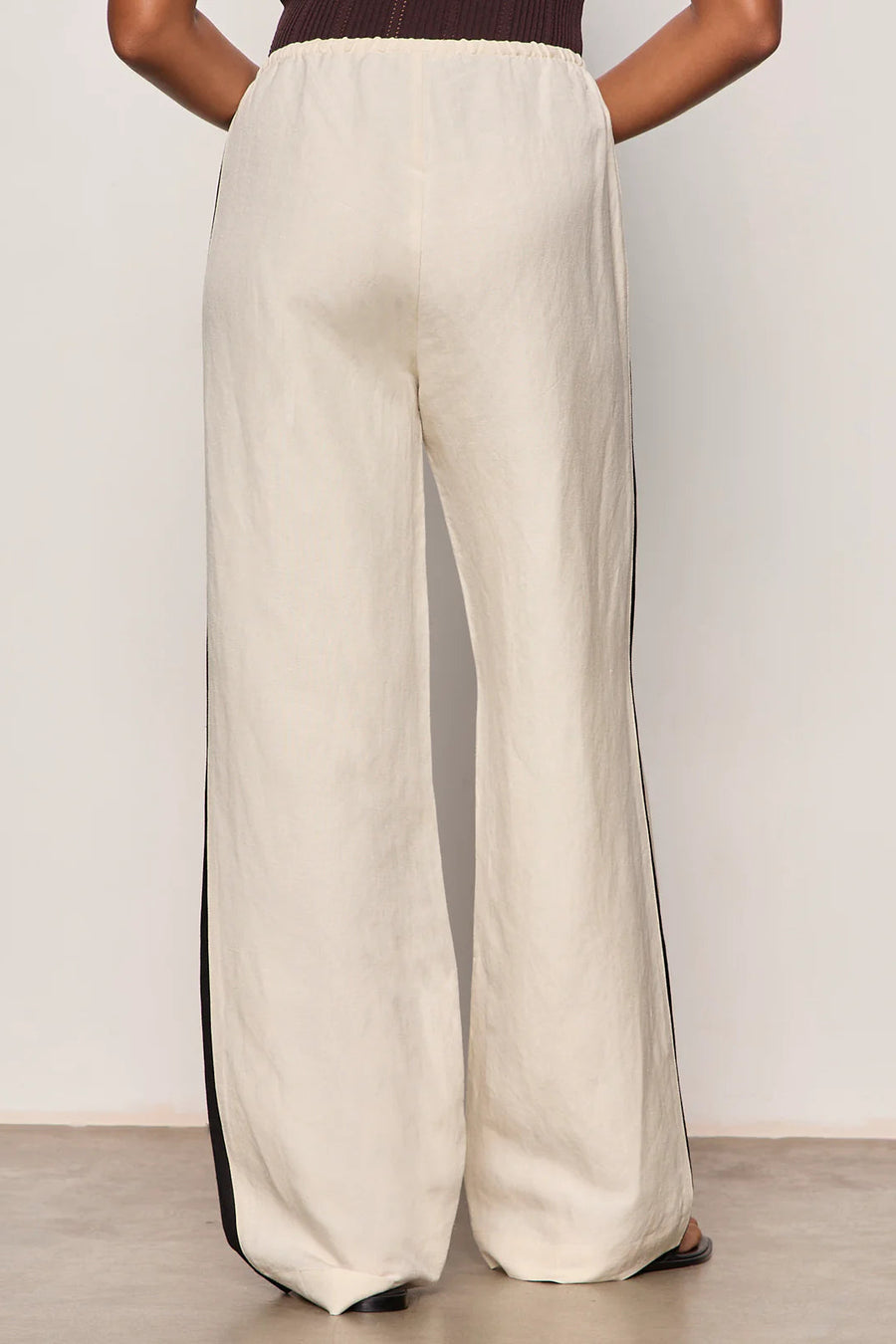 Woman wearing lightweight sand-colored wide-leg pants with contrasting black side stripe detailing. Features a comfortable pull-on elastic waistband, relaxed fit through the leg, and a fluid full-length silhouette. Designed with side pockets and a soft drape for effortless everyday wear, back