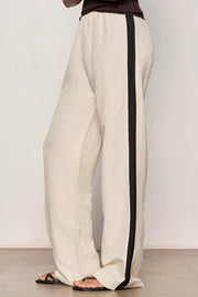 Woman wearing lightweight sand-colored wide-leg pants with contrasting black side stripe detailing. Features a comfortable pull-on elastic waistband, relaxed fit through the leg, and a fluid full-length silhouette. Designed with side pockets and a soft drape for effortless everyday wear, side